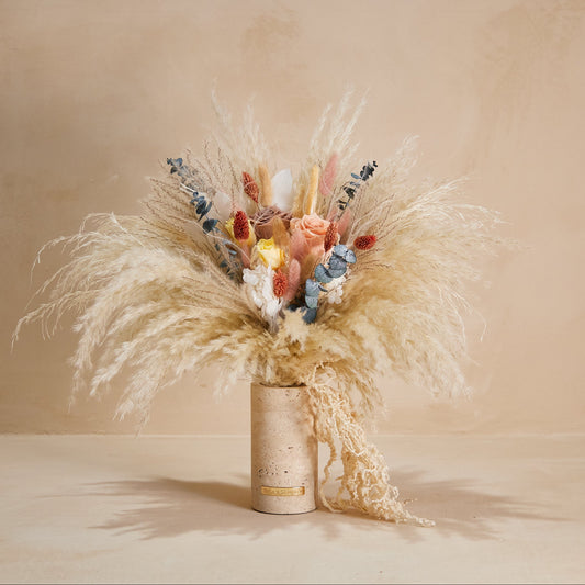 A dried floral arrangement featuring tall beige pampas grass, peach roses, blue eucalyptus leaves, and small yellow accents, displayed in a cylindrical beige vase against a warm beige background.