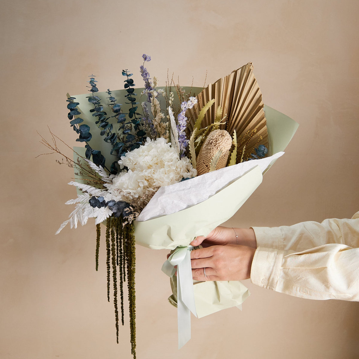 A dried flower bouquet with preserved hydrangeas and amaranthus wrapped in pale sage green paper